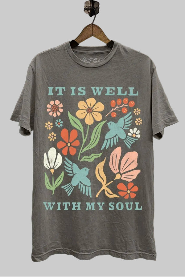 Gray t-shirt with colorful floral and bird design and 'It is well with my soul' text, hanging on a brick wall.