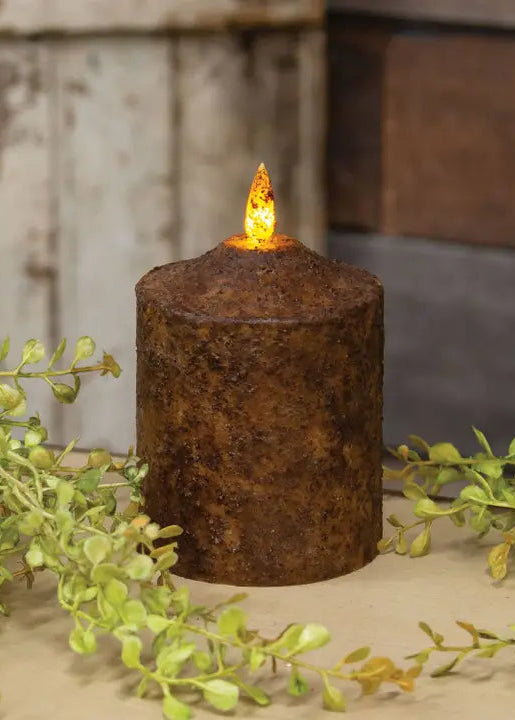 A rustic brown faux flameless LED pillar candle with a glowing orange flame effect, styled with green foliage on a neutral surface against a weathered wood background.