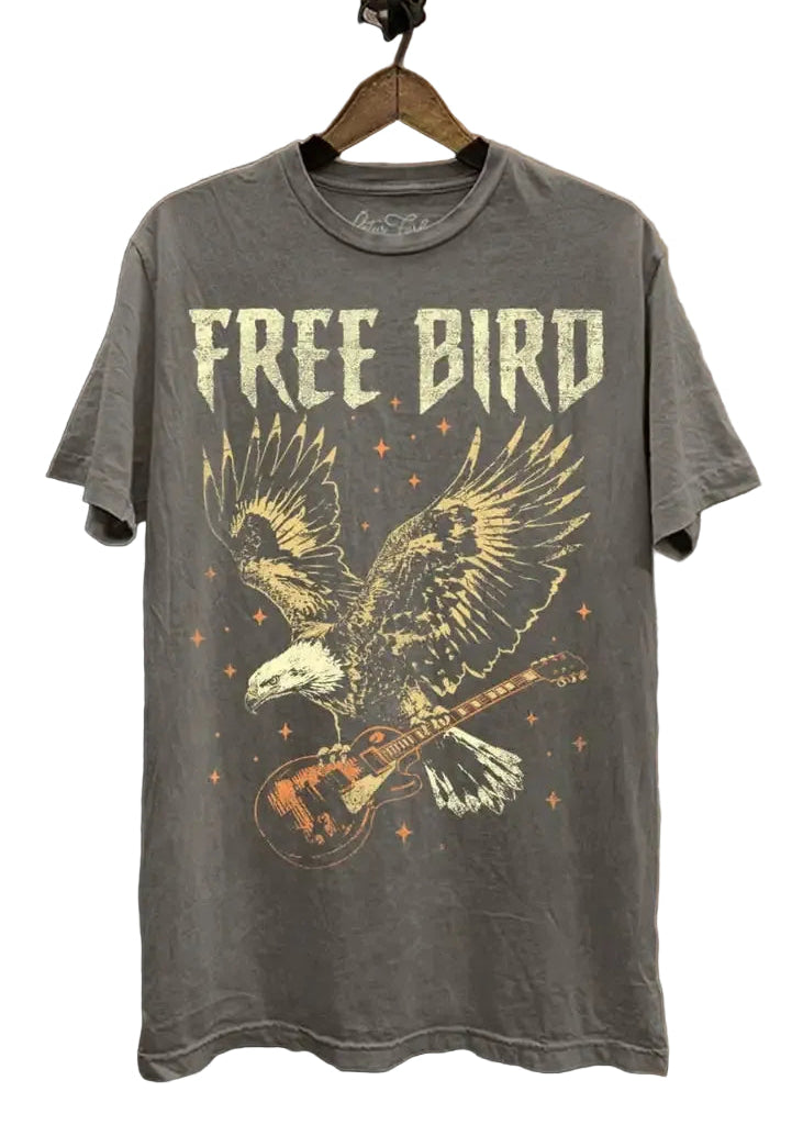 A gray graphic t-shirt hanging on a wooden hanger against a rustic brick and concrete wall. The shirt features a distressed graphic of a bald eagle playing an electric guitar with the words 'FREE BIRD' printed above it.