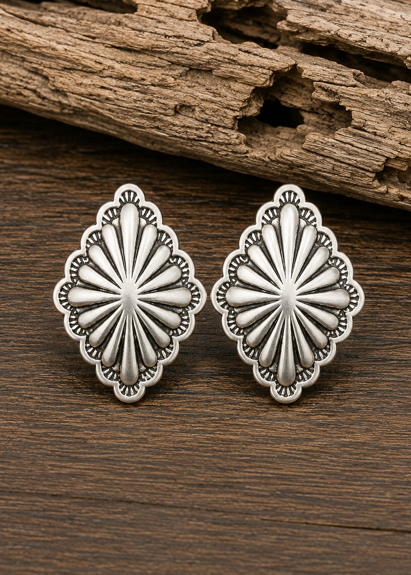 A pair of silver-toned concho-style stud earrings with a sunburst center and scalloped edges, displayed on a dark wood surface with a piece of weathered driftwood in the background.