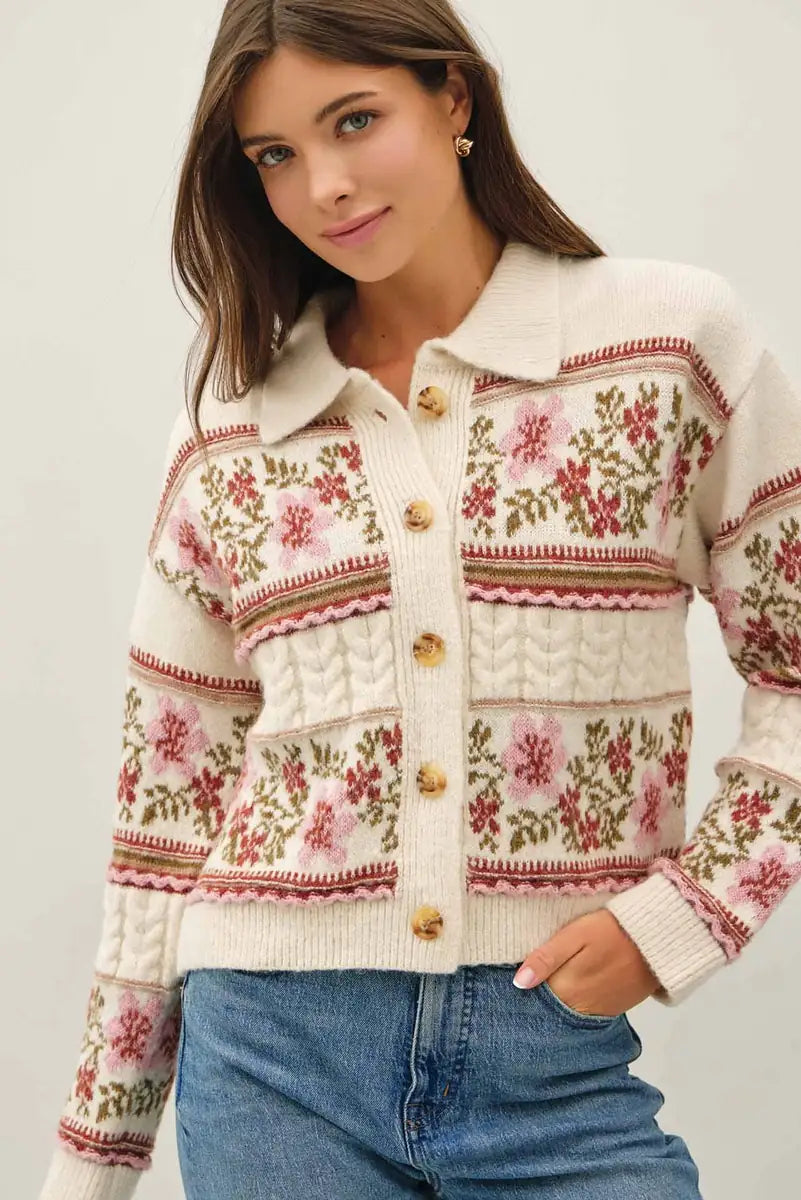 A woman wearing a cream-colored knit cardigan with pink floral patterns and brown buttons, paired with blue jeans, against a light gray background.