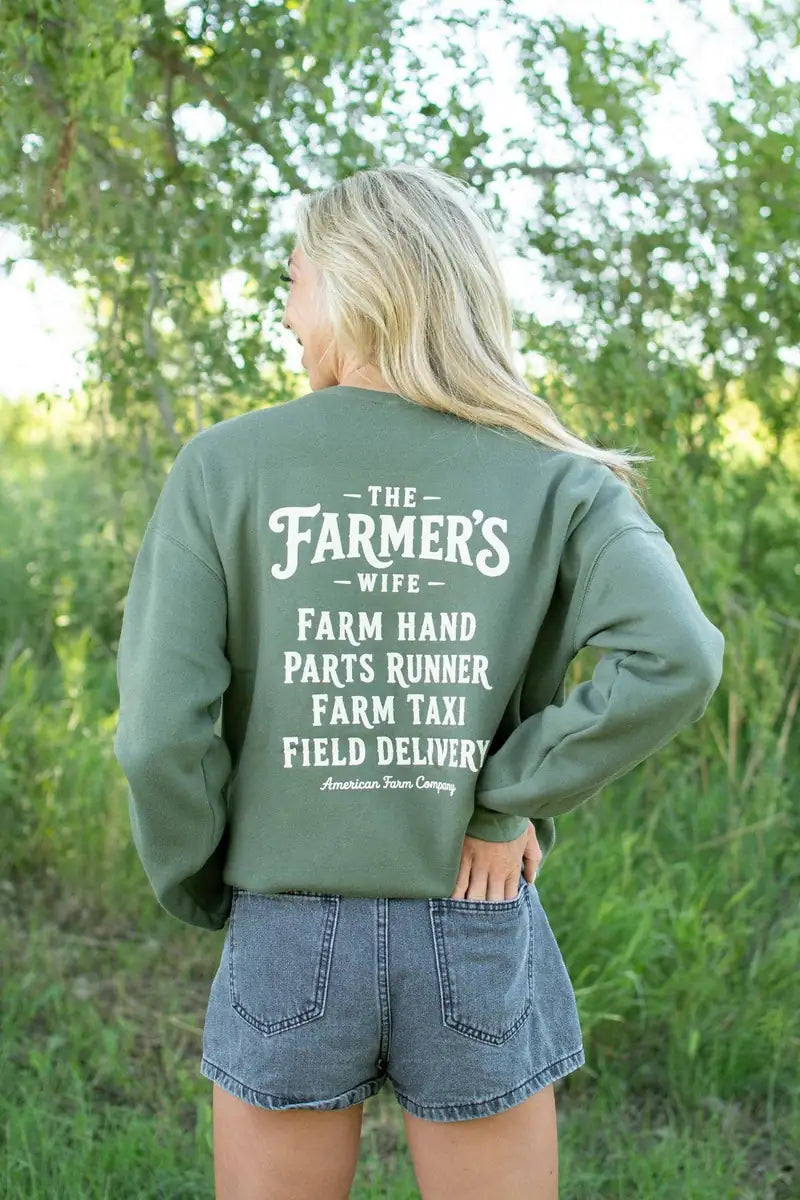 A woman viewed from behind wearing an olive green sweatshirt with white text that reads 'THE FARMER'S WIFE' and various farm-related job phrases, paired with denim shorts in an outdoor grassy setting.