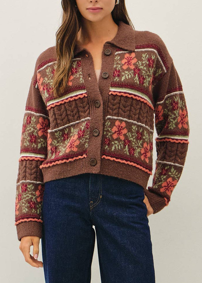A woman wearing a brown knit cardigan with floral and cable patterns in rust red, beige, and green, paired with dark blue jeans against a plain light gray background.