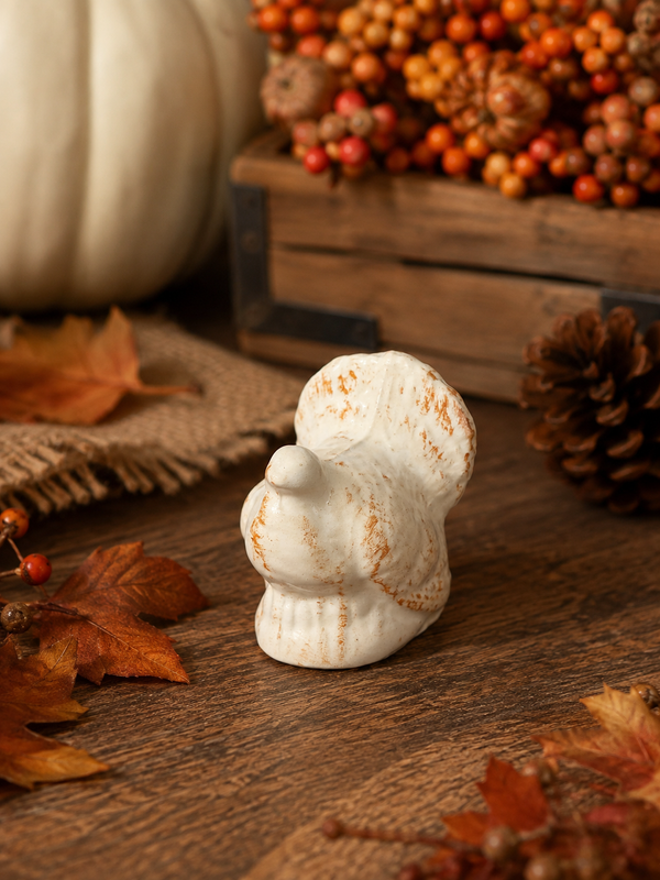 A small white ceramic decorative figurine with a rustic orange-brown distressed finish, styled on a wooden surface with autumn leaves, a pinecone, burlap, and a pumpkin in the background.