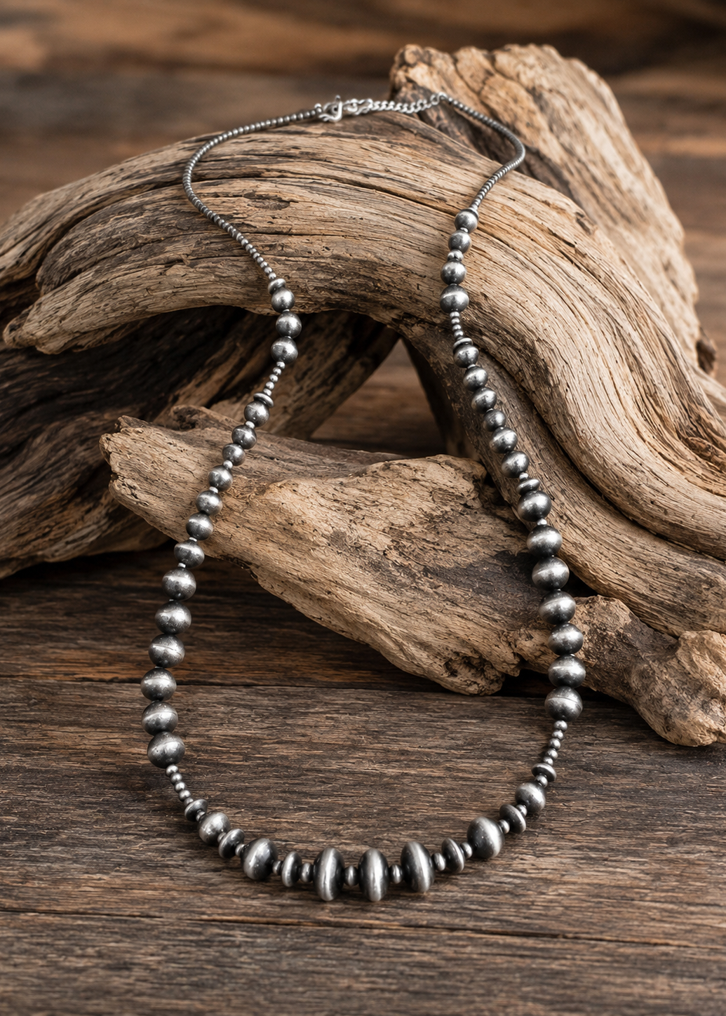 A beaded necklace made of dark metallic beads, draped over a piece of driftwood on a rustic wooden surface.