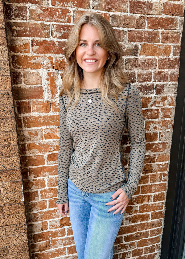 A woman with blonde hair stands smiling in front of a red brick wall, wearing a long-sleeve beige knit top with a small dark geometric pattern and light blue jeans.