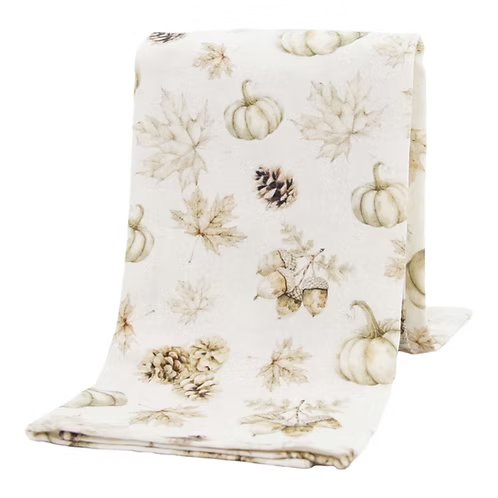 A folded fabric throw blanket with a light cream background and a printed autumn pattern featuring pumpkins, maple leaves, pinecones, acorns, and leaves in muted beige and brown tones.