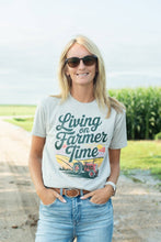Load image into Gallery viewer, Woman wearing a t-shirt with 'Living on Farmer Time' design in a rural setting.
