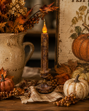 Load image into Gallery viewer, A rustic, dark brown faux-wax pillar candle with a glowing amber flame tip, styled on a wooden table with autumn decorations including pumpkins, a ceramic pitcher with fall foliage, and a plaid cloth.
