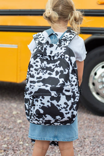 Child wearing a cow print backpack in front of a yellow school bus