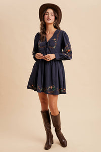 Woman wearing a navy dress with floral embroidery, brown boots, and a hat on a beige background