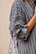 Load image into Gallery viewer, Close-up of a person wearing a black and white striped shirt with rolled-up sleeves against a beige background.