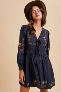 Woman wearing a navy dress with floral embroidery and a brown hat against a beige background