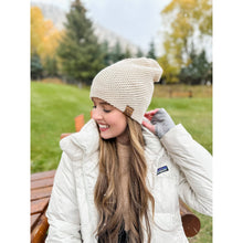 Load image into Gallery viewer, Woman wearing a beige knit beanie and white jacket in an outdoor setting with trees and grass.
