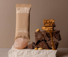 Load image into Gallery viewer, Chocolate bars with caramel and peanuts on a stone surface next to a branded packaging against a beige background
