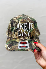 Load image into Gallery viewer, Camouflage cap with &#39;Lake It Easy&#39; text held by a hand against a gray background
