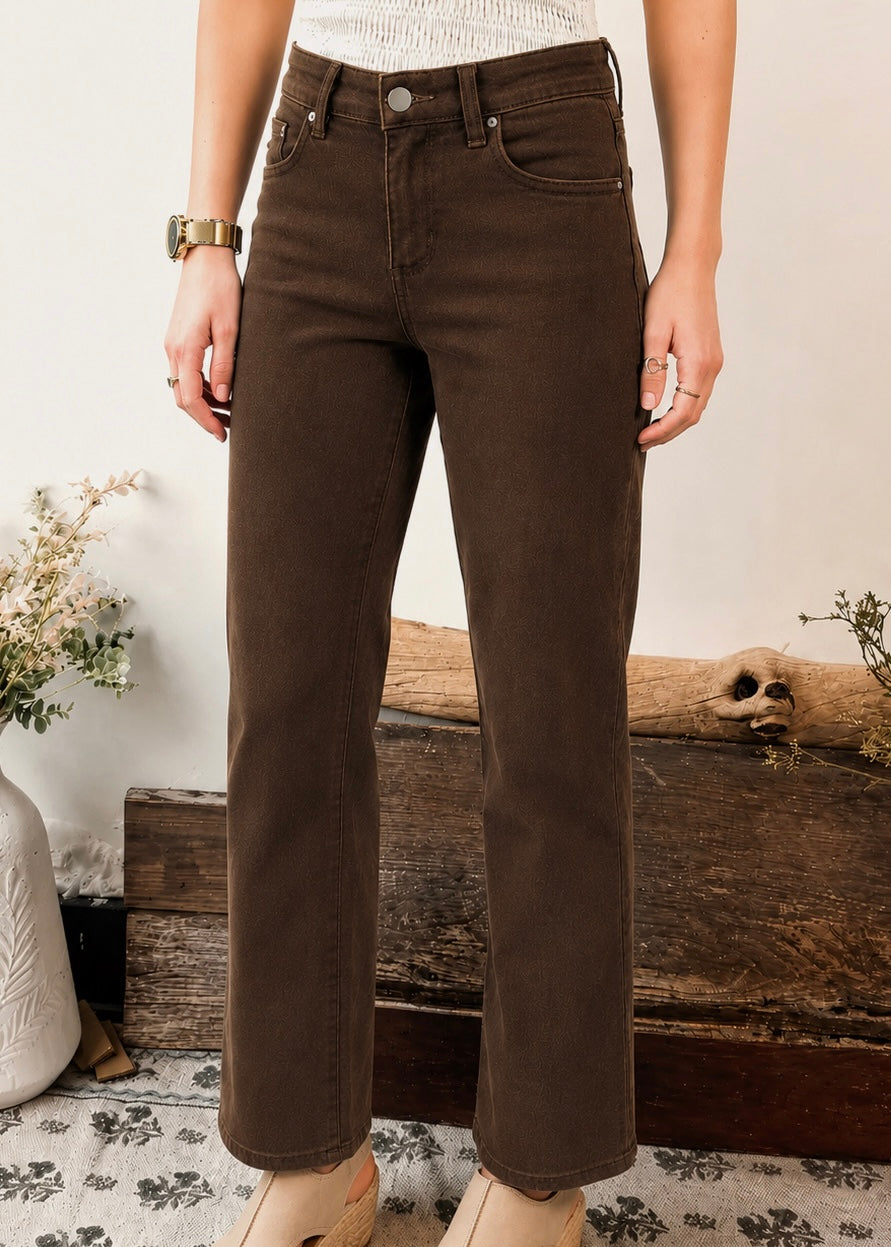 A person wearing dark brown straight-leg jeans, standing in front of a rustic wooden bench with dried flowers and a skull decor in a styled indoor setting.