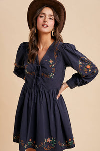 Woman wearing a navy dress with floral embroidery and a brown hat on a beige background