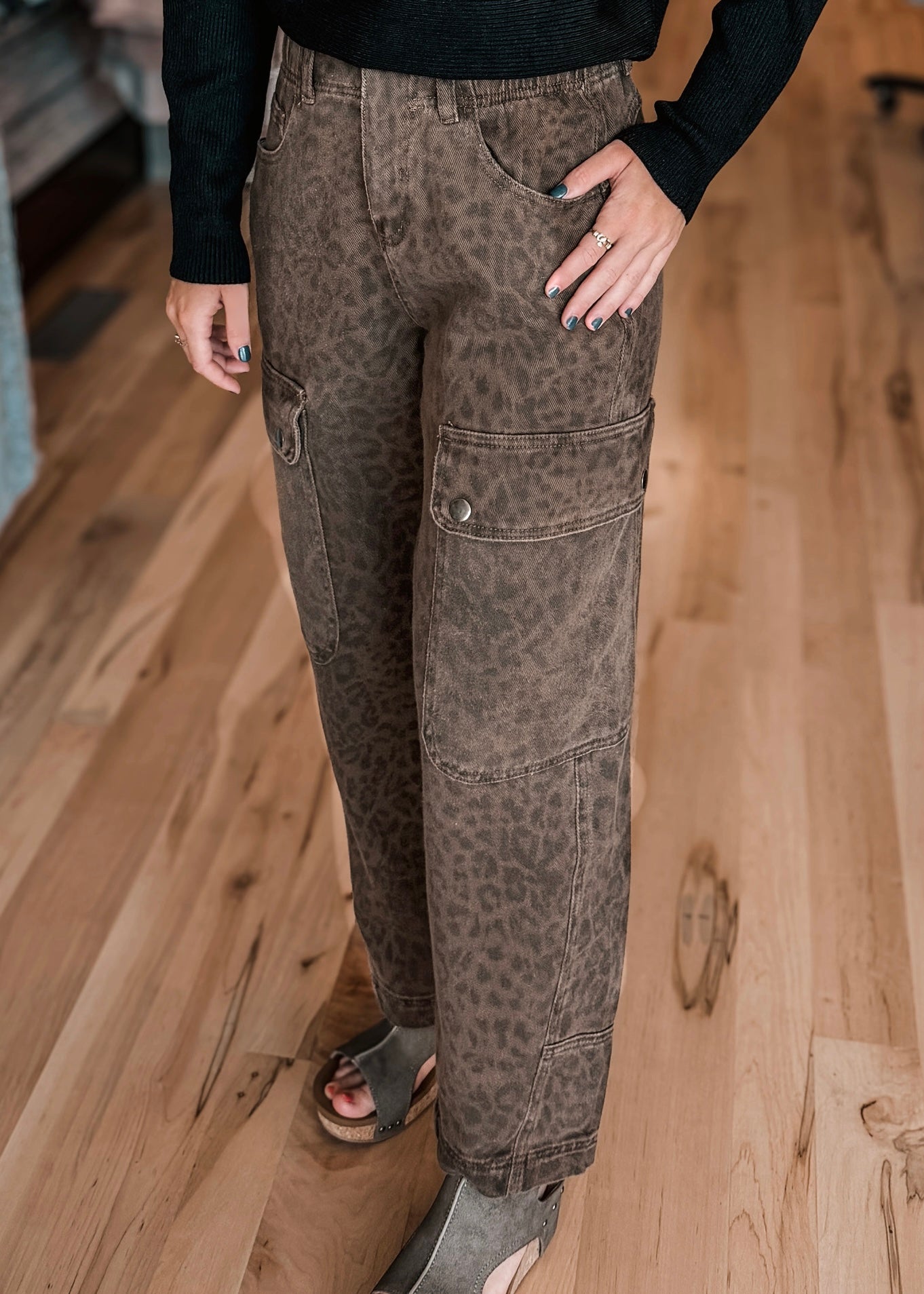 A person wearing brown leopard-print cargo pants with a black long-sleeve top and gray slide sandals, standing on a light wood floor.