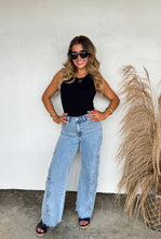 Load image into Gallery viewer, Woman wearing a black top and light blue jeans standing against a white wall with a plant to her right.