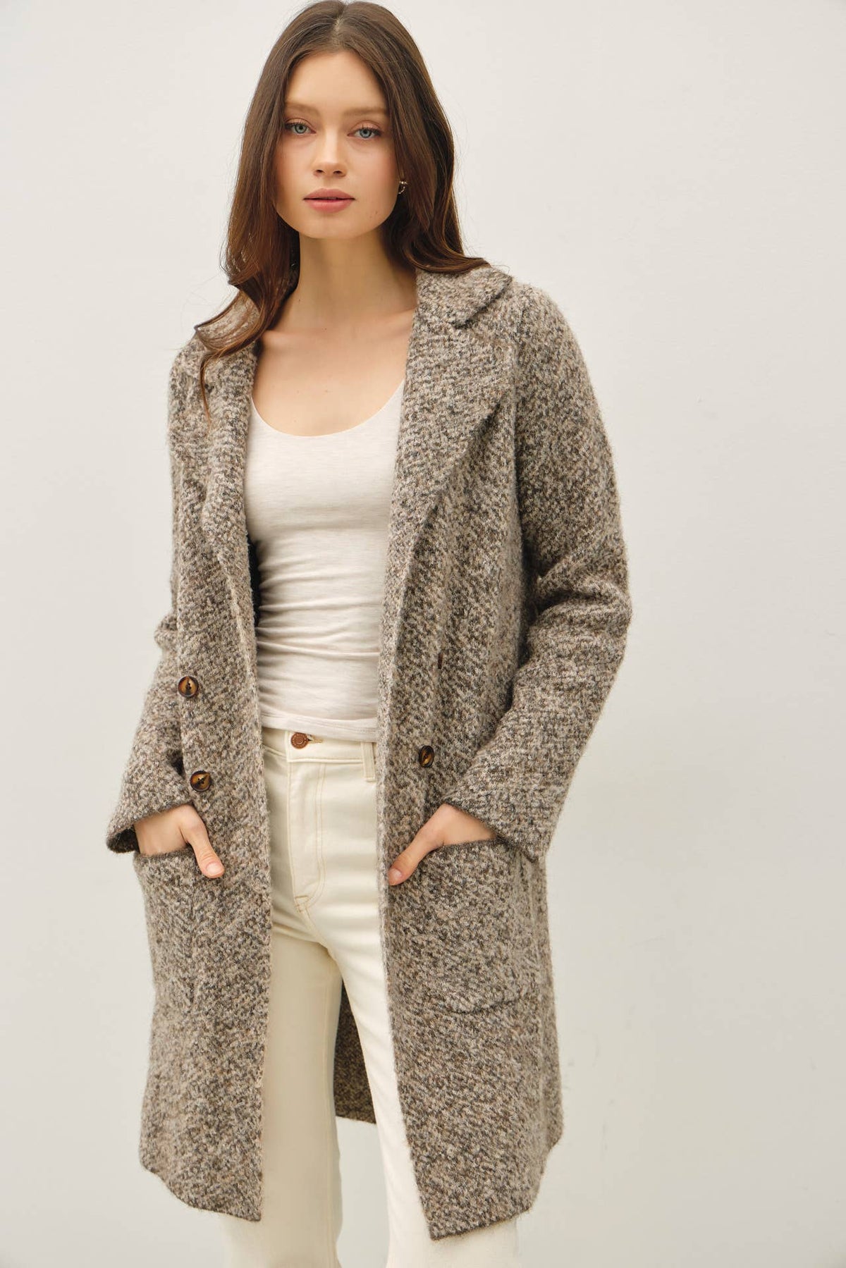Female model wearing a brown bouclé knit coatigan with a notched collar, double-button closure, long sleeves, and front patch pockets, styled with a cream tank and light-colored pants.