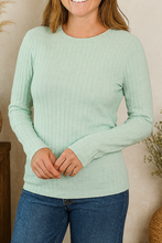 Load image into Gallery viewer, Woman wearing a light green sweater and blue jeans standing in a room with decorative plants.