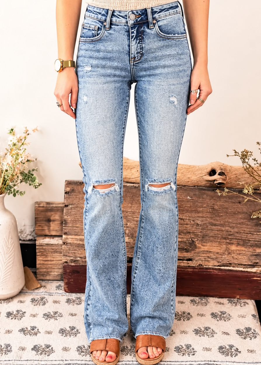 A person wearing light-wash blue distressed flare jeans with ripped knees, paired with a beige knit top and tan sandals, standing indoors in front of a rustic wooden chest and patterned rug.