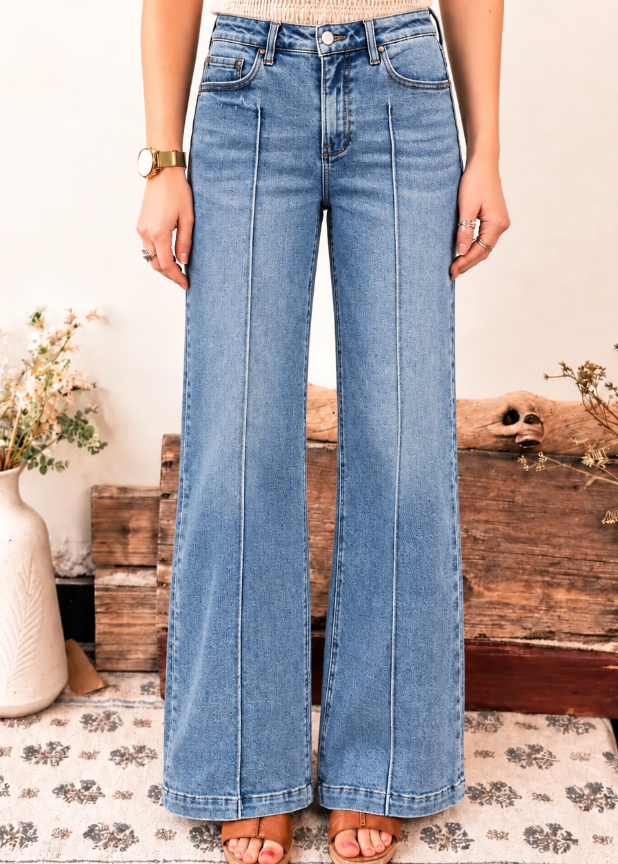 A person wearing light blue wide-leg denim jeans, styled with a beige knit top and tan sandals, standing in a rustic room setting with wooden crates and dried flowers.