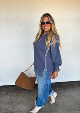 Load image into Gallery viewer, Woman wearing a blue shirt and jeans, holding a brown bag against a white wall.