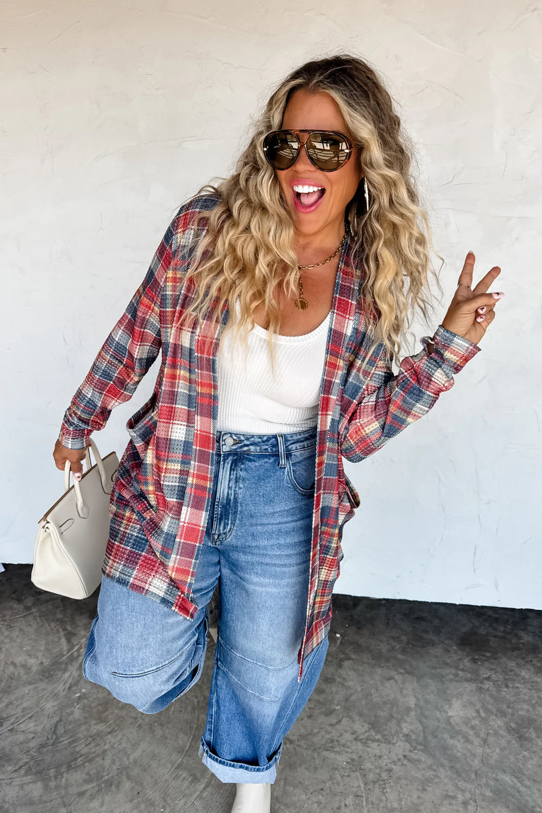Woman wearing a plaid shirt, white top, and jeans, posing against a white wall.