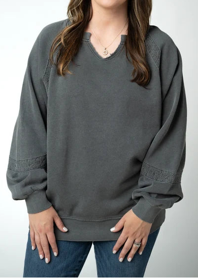 A woman with long brown hair and bangs smiling while wearing a dark gray v-neck sweatshirt with lace-up details on the sleeves, paired with blue jeans, against a light gray studio background.