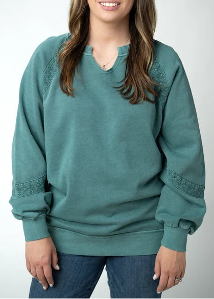 A woman with long brown hair and bangs smiling while wearing a teal green v-neck sweatshirt with lace-up details on the sleeves, paired with blue jeans, against a light gray studio background.