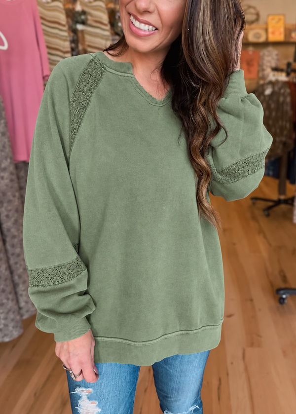 A woman wearing an olive green crewneck sweatshirt with textured lace-like trim on the shoulders and cuffs, paired with distressed blue jeans in a clothing boutique setting.
