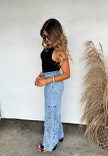 Load image into Gallery viewer, Woman wearing a black top and light blue pants against a white wall with decorative elements.