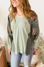 Load image into Gallery viewer, Woman wearing a green floral-patterned top and jeans standing in a home setting with decorative elements.