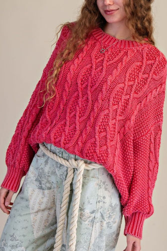 Person wearing a pink cable knit sweater and light-colored pants against a neutral background