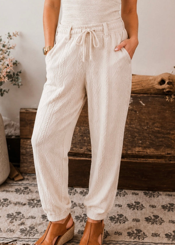 A person wearing cream-colored textured drawstring pants with side pockets, paired with tan wedge sandals, standing in a rustic indoor setting.