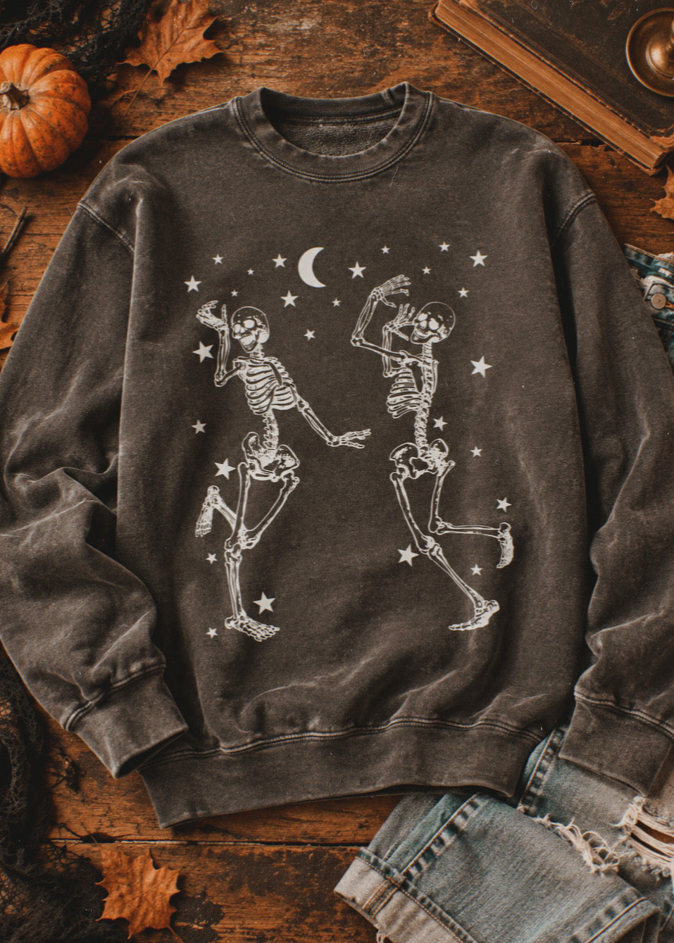 A dark gray acid-wash crewneck sweatshirt featuring a white graphic of two dancing skeletons under a crescent moon and stars, styled in a flat lay on a rustic wooden table with autumn leaves, a pumpkin, and denim jeans.