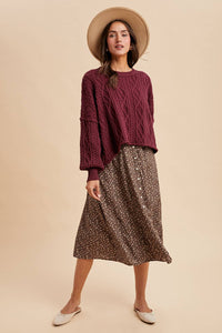Woman wearing a burgundy cable knit sweater and patterned skirt with a beige hat on a beige background