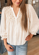 Load image into Gallery viewer, A woman wearing a cream-colored textured blouse with a geometric embroidered yoke and puff sleeves, paired with light-wash blue jeans in a clothing store setting.
