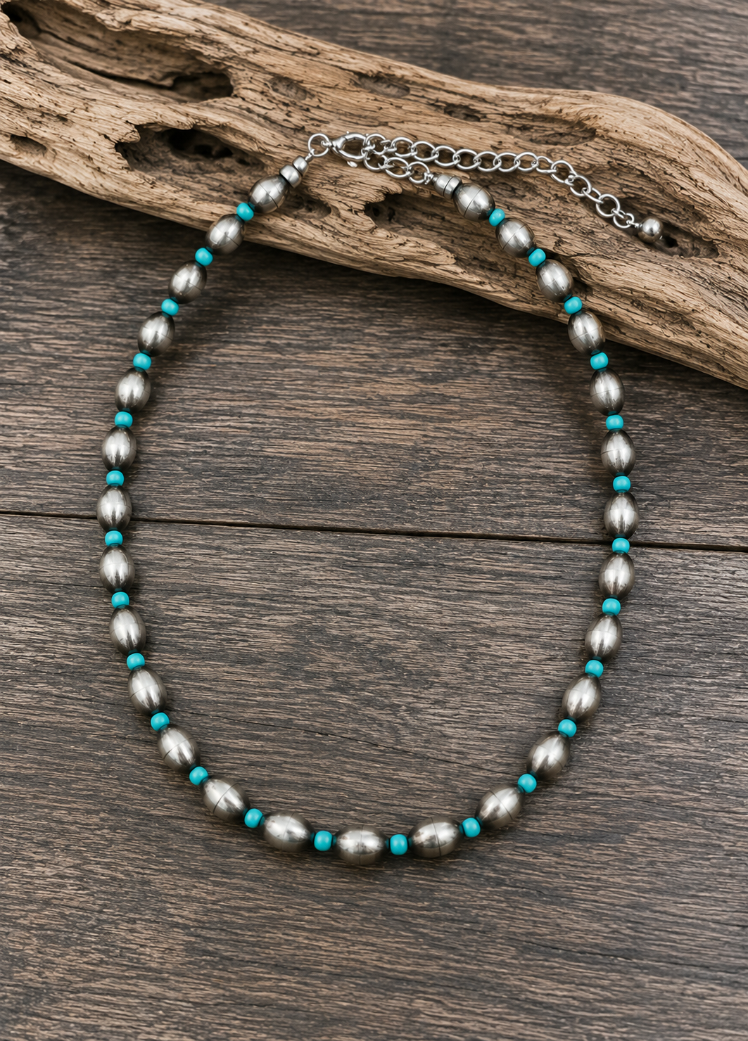 A beaded necklace with metallic silver-gray oval beads and small turquoise blue spacer beads, displayed on a rustic wood surface with a piece of driftwood.