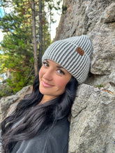 Load image into Gallery viewer, Woman wearing a striped beanie leaning against a rock wall with trees in the background