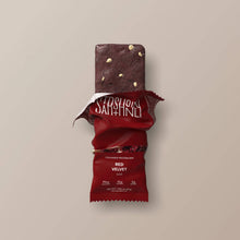 Load image into Gallery viewer, Red velvet protein bar with a partially eaten wrapper on a beige background