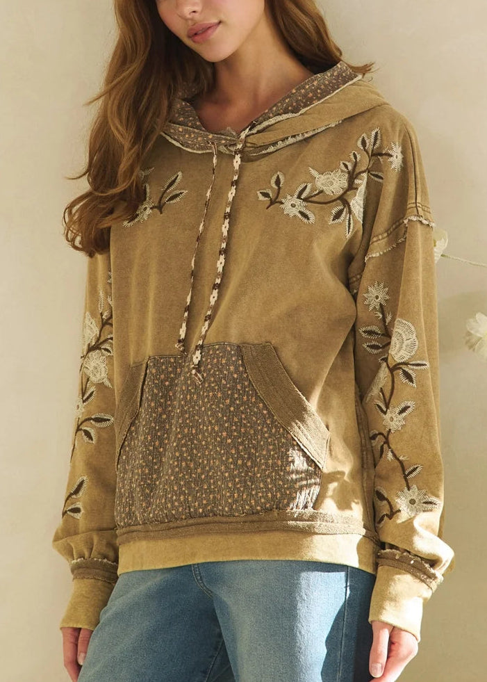 A woman wearing a tan embroidered hoodie with floral patterns and a patterned front pocket, paired with blue jeans, standing against a neutral beige wall.