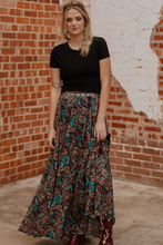 Load image into Gallery viewer, Woman wearing a black top and patterned skirt standing against a brick wall.